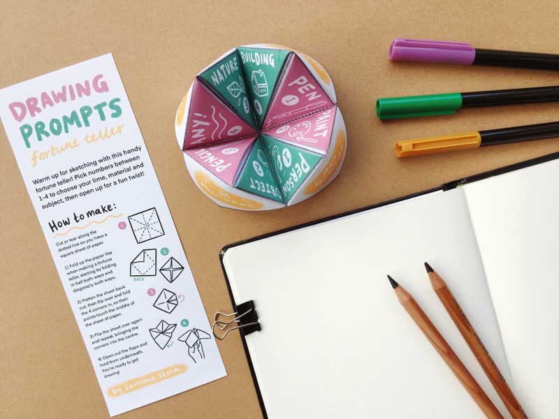 An open sketchbook with pencils resting on it, next to three colourful marker pens. A folded paper fortune teller sits next to it, open with green and pink sections inside.